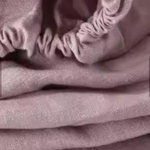 Op de afbeelding: Een kledingstuk van oudroze stof met een geplooide, elastische opening aan de bovenkant. Het materiaal heeft een zachte, matte textuur met zichtbare plooien en draperingen, wat zorgt voor een gelaagd uiterlijk over het textieloppervlak.