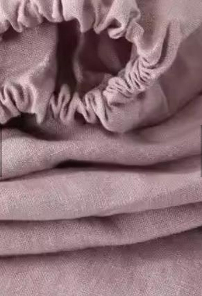 Op de afbeelding: Een kledingstuk van oudroze stof met een geplooide, elastische opening aan de bovenkant. Het materiaal heeft een zachte, matte textuur met zichtbare plooien en draperingen, wat zorgt voor een gelaagd uiterlijk over het textieloppervlak.