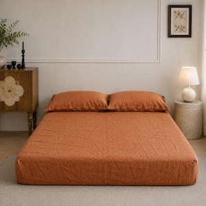 Op de afbeelding: Een bed met een effen roestoranje hoeslaken en twee bijpassende rechthoekige kussens. Het beddengoed heeft een zichtbaar gestructureerd stoffen oppervlak en is netjes op een laag bedframe gerangschikt, wat zorgt voor een uniforme kleurstelling.