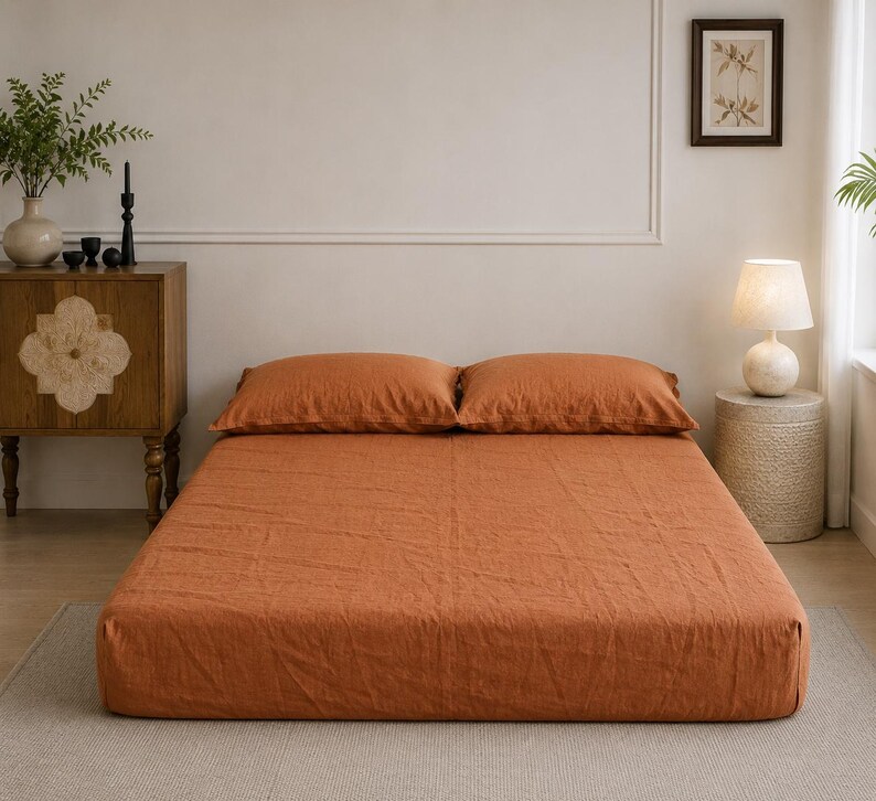Op de afbeelding: Een bed met een effen roestoranje hoeslaken en twee bijpassende rechthoekige kussens. Het beddengoed heeft een zichtbaar gestructureerd stoffen oppervlak en is netjes op een laag bedframe gerangschikt, wat zorgt voor een uniforme kleurstelling.