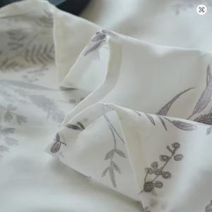 May include: A white fabric featuring a repeating botanical print in a muted grey tone. The pattern consists of various delicate leaves, stems, and small berries scattered across the surface. The material is folded, creating soft, layered pleats.