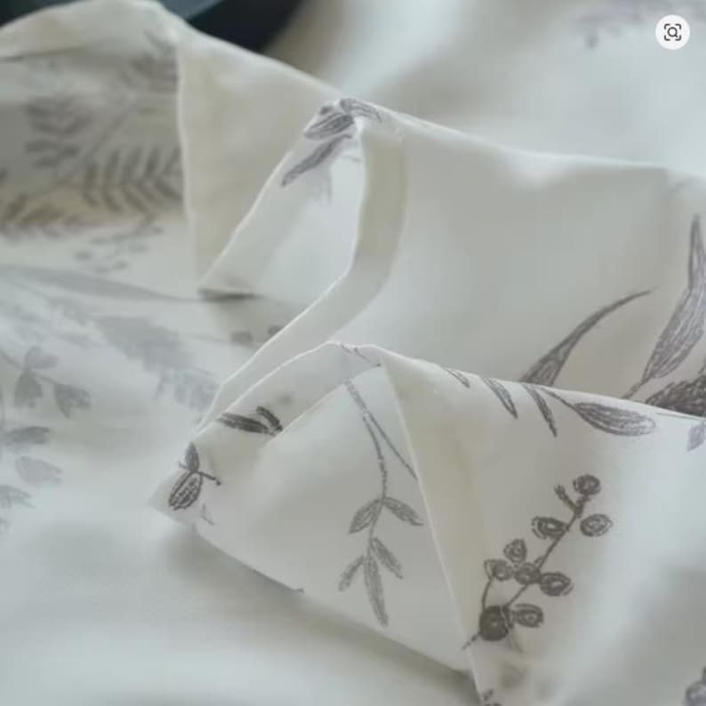 May include: A white fabric featuring a repeating botanical print in a muted grey tone. The pattern consists of various delicate leaves, stems, and small berries scattered across the surface. The material is folded, creating soft, layered pleats.