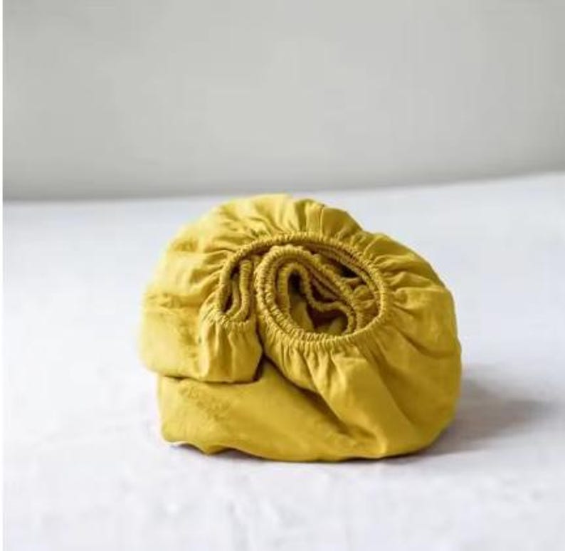 Op de afbeelding: Een mosterdgeel hoeslaken met een elastische rand, opgevouwen in een cirkelvorm. De stof heeft een zichtbare textuur en een matte afwerking, waarbij de geplooide elastische band langs de omtrek van het textiele beddengoedartikel zichtbaar is.