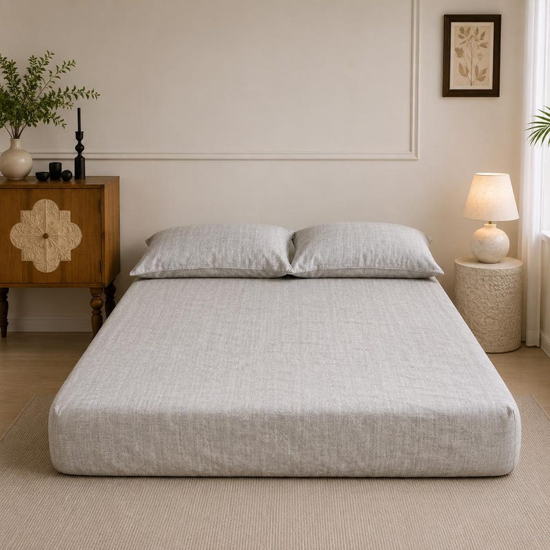 Op de afbeelding: Een bed met een lichtgrijs, gestructureerd hoeslaken in linnenlook en twee bijpassende rechthoekige kussens. Het beddengoed heeft een subtiele, geweven uitstraling met een neutrale tint, wat zorgt voor een strak en uniform oppervlak.