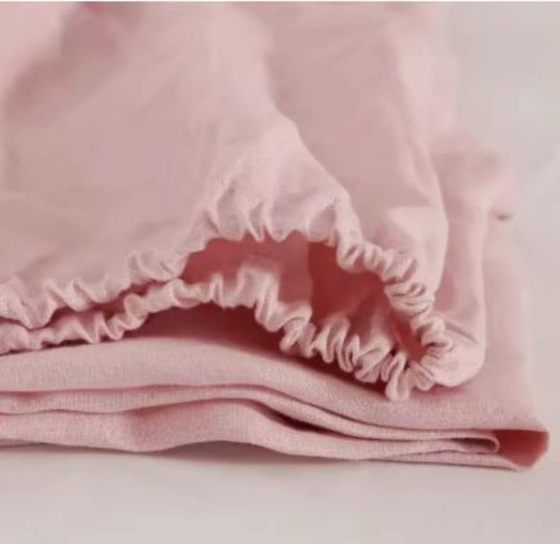 Op de afbeelding: Een lichtroze stoffen laken met een geplooide, elastische rand. Het materiaal heeft een zachte, geweven textuur met zichtbare vouwen en kreukels, wat de flexibele en rekbare aard van de gezoomde rand voor een goede pasvorm benadrukt.