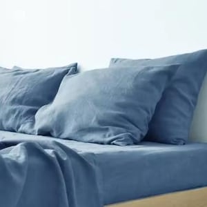 Op de afbeelding: Een set beddengoed in een stoffige blauwe kleur, bestaande uit twee rechthoekige kussens en een bijpassend hoeslaken. De stof heeft een zichtbaar, licht getextureerd weefsel en een matte afwerking, liggend op een matras met een licht houten frame.