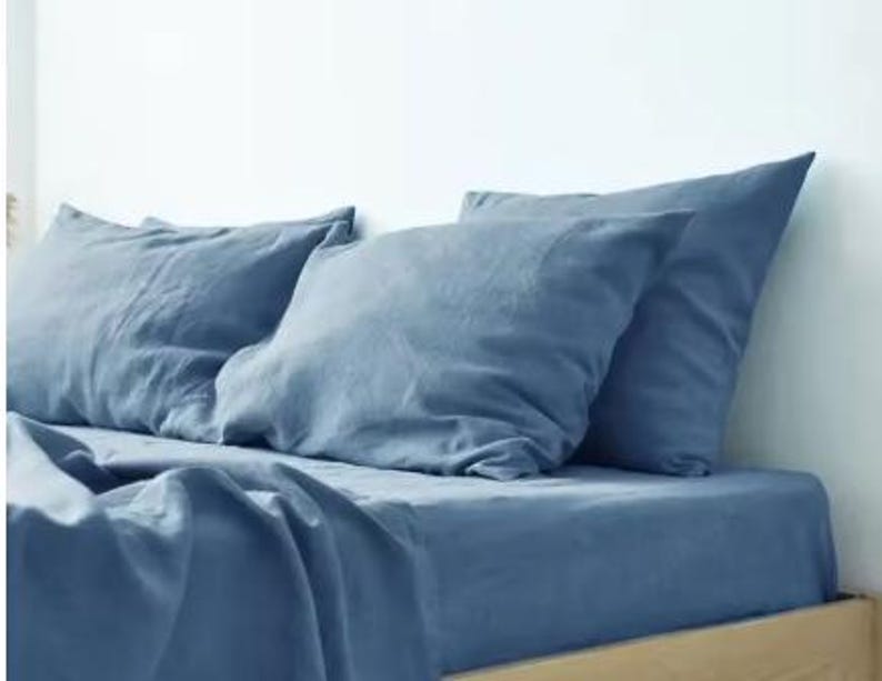 Op de afbeelding: Een set beddengoed in een stoffige blauwe kleur, bestaande uit twee rechthoekige kussens en een bijpassend hoeslaken. De stof heeft een zichtbaar, licht getextureerd weefsel en een matte afwerking, liggend op een matras met een licht houten frame.