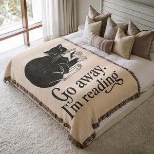 May include: A beige throw blanket featuring a black cat illustration and the text "Go away. I'm reading." The blanket has a fringed edge and is placed on a bed with pillows. The cat is shown holding a book.