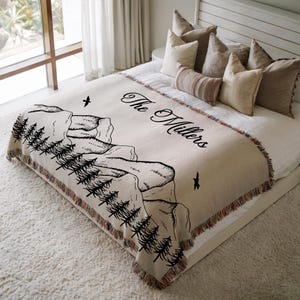 May include: Beige woven throw blanket with a black mountain and tree design. The blanket has a fringed edge and the text "The Millers" in a script font. The blanket is on a bed with pillows.