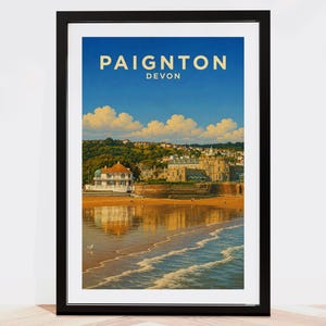 Pode incluir: Um cartaz de viagem emoldurado apresentando uma cena costeira em Paignton, Devon. A imagem exibe uma praia de areia com ondas suaves, um grande edifício com fachada branca e um céu azul claro com nuvens. O texto PAIGNTON DEVON está no topo.
