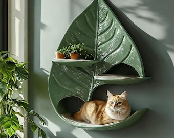 Può includere: Una mensola da parete per gatti a forma di grande foglia verde con finitura lucida. Presenta due piattaforme superiori e una base inferiore più ampia, ciascuna rivestita con un cuscinetto in tessuto chiaro. Un gatto rosso riposa in basso.