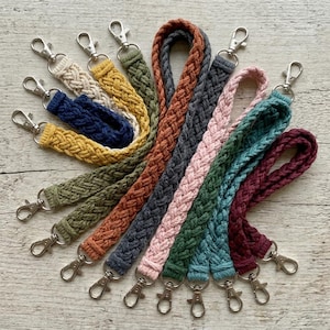 Könnte beinhalten: Eine Sammlung geflochtener Handgelenk-Lanyards in verschiedenen Farben, darunter Beige, Gelb, Marineblau, Olivgrün, Orange, Grau, Rosa, Türkis und Burgunderrot. Jedes Lanyard hat einen silberfarbenen Karabinerhaken zum Befestigen von Schlüsseln oder anderen Gegenständen.