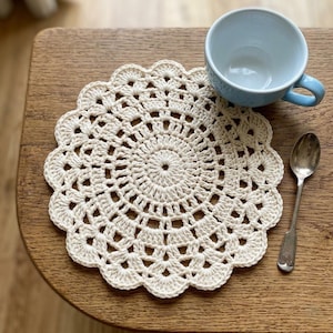 May include: A cream-colored, crocheted doily with a scalloped edge. A light blue teacup and a silver spoon rest on a wooden surface next to the doily. The doily has a circular design with a central spiral pattern.