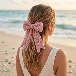 Op de afbeelding: Een grote, stoffige roze gehaakte haaraccessoire. De strik is bevestigd aan de achterkant van het hoofd van een persoon, met de oceaan en het strand op de achtergrond. De strik is gemaakt van een getextureerd garen en heeft lange, vloeiende staarten.