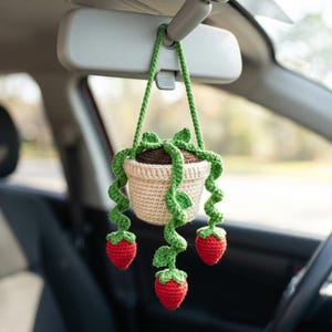 May include: A crocheted car charm featuring a beige pot with green vines and red strawberries. The pot is filled with brown material, and the vines have green leaves. The charm hangs from a green cord attached to a car's rearview mirror.