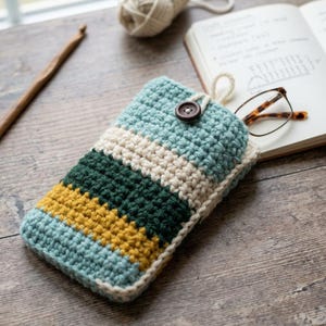 May include: A handmade crochet glasses case in shades of blue, cream, green, and yellow. The case features a button closure and a loop. A pair of eyeglasses rests on a notebook next to the case. A ball of yarn and a wooden crochet hook are also visible.