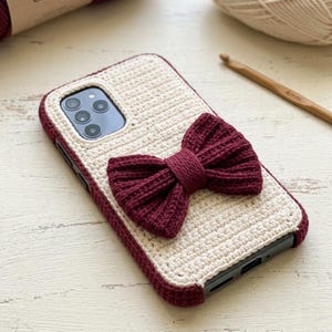 May include: Cream-colored crochet phone case with a burgundy bow. The case has a burgundy border and is designed to fit a smartphone. The bow is centered on the case and adds a decorative touch. The case is handmade and likely crafted from yarn.