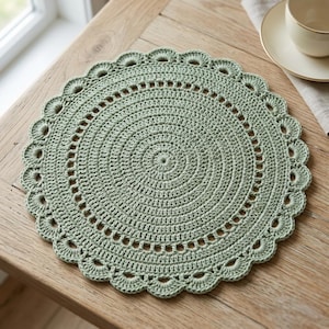 May include: A sage green, crocheted placemat with a scalloped edge. The circular mat features concentric circles and decorative openwork. The placemat is on a wooden table, with a teacup and saucer visible in the background.