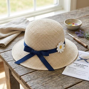 May include: A beige crochet sun hat with a dark blue ribbon and a white daisy accent. The hat has a wide brim and a rounded crown. The hat is sitting on a wooden surface.
