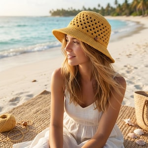 May include: A yellow crochet sun hat with a wide brim and a wooden button detail. The hat is worn by a person sitting on a beach, with the ocean and palm trees in the background. The hat is made of a textured yarn.