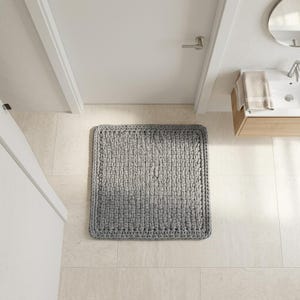 May include: A rectangular, gray bath mat with a thick, woven texture. The mat is placed on a light-colored tiled floor in a bathroom setting. The mat's dimensions are approximately 24 x 36 inches.