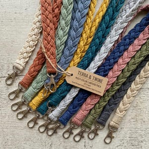 Könnte beinhalten: Eine Sammlung mehrfarbiger geflochtener Baumwoll-Lanyards mit Edelstahlbeschlägen. Die Lanyards sind in verschiedenen Farben erhältlich, darunter Beige, Orange, Grün, Blau und Grau. Ein Etikett lautet "TERRA & TWINE MULTI-COLOR LANYARD PACK (12 Straps)".