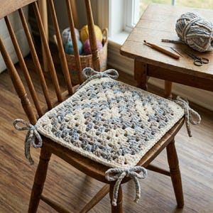 May include: A handmade, square crochet chair cushion in cream, blue, and gray tones, tied to the chair with matching yarn bows. The cushion sits on a wooden chair with a spindle back. A small table holds yarn, a crochet hook, and scissors.