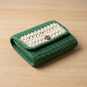 May include: A handmade green crochet wallet with a cream-colored decorative flap. The wallet is rectangular with a magnetic snap closure. The crochet stitch is visible, and the wallet is on a wooden surface.