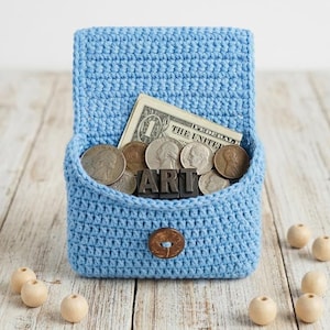 Puede incluir: Un bolso de ganchillo azul claro, lleno de monedas, un billete de dólar y la palabra "ART" en letras metálicas. El bolso se cierra con un botón marrón. Hay cuentas de madera esparcidas.