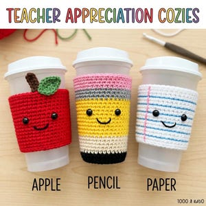 May include: Three white plastic cups with crocheted cozies. One is red with an apple design, one is yellow, gray, and pink like a pencil, and one is white with blue lines like notebook paper. Each cozy features a smiling face with black eyes.