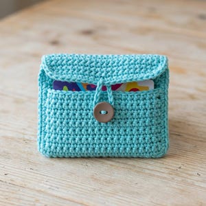 May include: A turquoise crocheted wallet with a flap closure and a brown button. The wallet has a pocket on the front and is made of a textured yarn. The wallet is designed to hold cards.