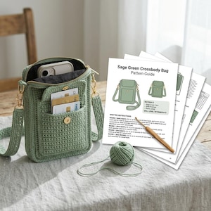 May include: A sage green crocheted crossbody bag with a front pocket containing credit cards, a smartphone inside, and a gold-tone snap closure. Beside it lies a ball of matching yarn, a wooden crochet hook, and a printed pattern guide titled Sage Green Crossbody Bag Pattern Guide.