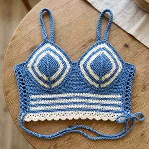 Peut inclure: Haut au crochet bleu et blanc avec un design bustier. Le haut présente un motif géométrique sur les bonnets et des rayures horizontales. Les bretelles et les liens sont bleus, et le bord inférieur a une bordure festonnée.
