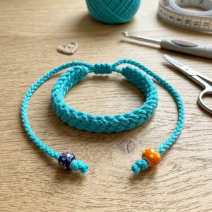 May include: A turquoise crochet bracelet with a braided design, featuring decorative beads at the ends. The bracelet is displayed on a wooden surface, alongside a ball of turquoise yarn, a crochet hook, scissors, and a measuring tape.