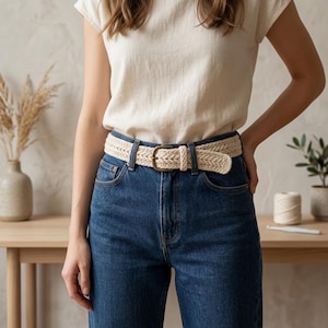 May include: A cream-colored woven belt featuring a braided texture and a simple metal buckle. The belt is worn through the belt loops of dark blue denim jeans, which are paired with a plain, short-sleeved cream-colored t-shirt.