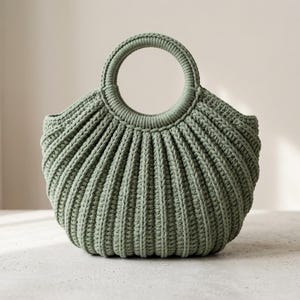 Puede incluir: Un bolso de ganchillo en color verde salvia con una textura acanalada distintiva y un asa circular. El bolso presenta una silueta redondeada en forma de concha con patrones de costura verticales que crean un aspecto estructurado.