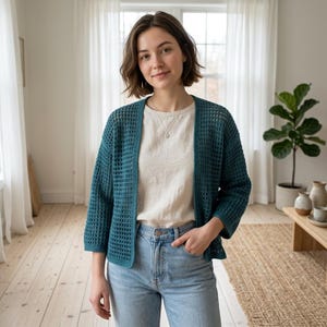 May include: A teal-colored, open-front cardigan with a waffle-knit pattern. The cardigan is worn over a cream-colored top and paired with light blue jeans. The outfit is completed with a silver necklace.