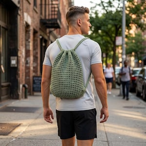May include: A green, open-weave drawstring backpack. The backpack is carried on the back. The bag has a rounded shape and is made of a textured material. The straps are a matching green color. The background shows a city street.