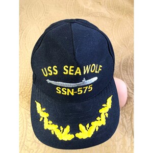 May include: A black baseball cap featuring yellow embroidered text that reads USS SEA WOLF and SSN-575. A silver embroidered submarine graphic is positioned between the text. The brim displays a yellow embroidered leaf-like pattern.