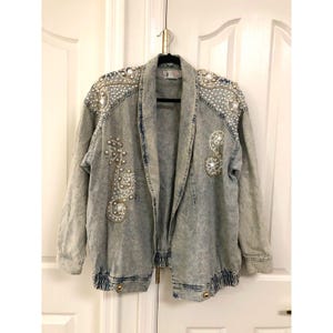 May include: An acid-wash blue denim jacket featuring a shawl collar, long sleeves, and an elasticized hem. The jacket is embellished with white faux pearls and gold-toned beads arranged in swirling patterns across the shoulders and front panels.