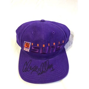May include: A purple baseball cap featuring the Phoenix Suns logo and text embroidered in orange and purple on the front crown. The brim displays a black handwritten signature across the center. The cap has a structured six-panel design.
