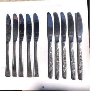 May include: A set of ten stainless steel dinner knives arranged in two rows of five. The five knives on the left feature a simple geometric pattern at the base of the handle, while the five knives on the right display an embossed floral design.