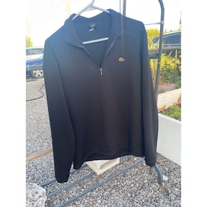 May include: Black long-sleeve pullover with a quarter-zip closure. The shirt has a small logo on the chest and is hanging on a metal hanger. The garment is made of a soft, dark fabric.