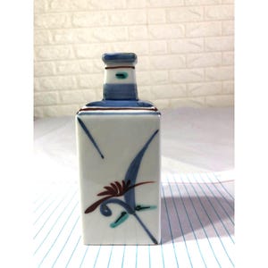 May include: A rectangular white ceramic bottle with a narrow neck, decorated with blue and maroon floral designs. The bottle's neck is blue with white and maroon accents. The bottle is on a white surface.