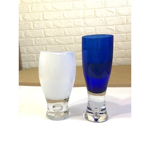 Könnte beinhalten: Zwei Glasvasen mit dicken, klaren Böden, die jeweils eine Luftblase enthalten. Eine Vase ist undurchsichtig weiß mit bauchigem Körper, die andere ist eine hohe, zylindrische Vase aus tiefblauem Glas. Beide haben glatte, polierte Oberflächen.