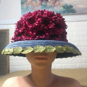 Könnte beinhalten: Ein breitkrempiger Hut mit einem einzigartigen Blumendesign. Die Krone ist mit burgunderfarbenen Blumen bedeckt, während die Krempe blau mit grünen Blättern verziert ist. Der Hut wird auf einer Schaufensterpuppe präsentiert.