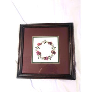 Pode incluir: Arte floral emoldurada quadrada. A obra apresenta uma coroa de flores roxas, rosas e azuis sobre um fundo branco, sobre uma passe-partout bordô. A moldura é marrom escuro.