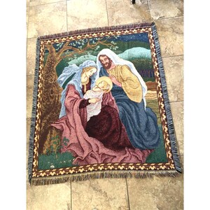 May include: A rectangular woven tapestry throw blanket featuring a nativity scene with a central figure holding a baby, surrounded by a figure in a blue robe and a large tree. The border has a repeating brown and tan geometric pattern with fringe.