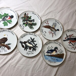 May include: A collection of seven white ceramic plates with gold rims, each featuring a unique bird illustration, the text Kempton's Travel Town, a specific year, and the text Jayco Inc. The birds include a bullfinch, cardinal, pheasant, eagle, chickadee, wood duck, and robin.