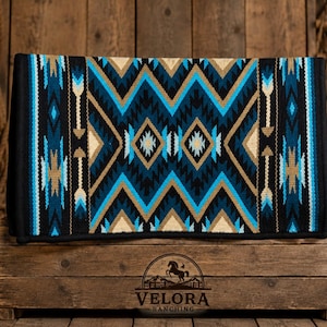 May include: A rectangular horse saddle pad featuring a geometric pattern in black, bright blue, and tan. The design includes repeating zigzags, diamonds, and arrow motifs. The pad has a solid black border and is displayed on a wooden crate.